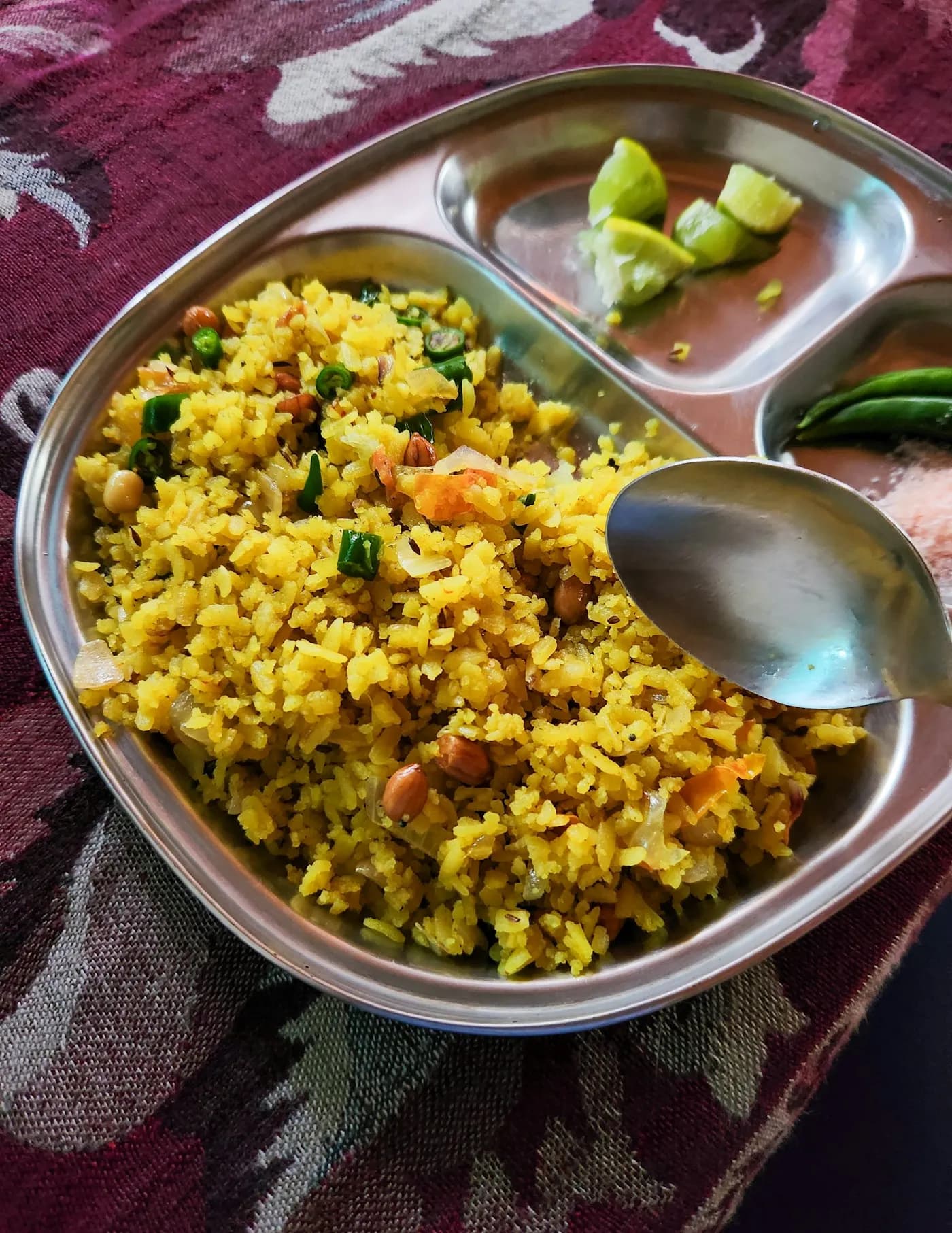 A steel plate of yellow poha with peanuts, green chillies and lime wedges